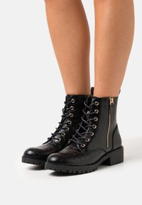 Bottines noires en simili cuir avec un motif crocodile, des détails dorés, un laçage et une fermeture éclair sur le côté.