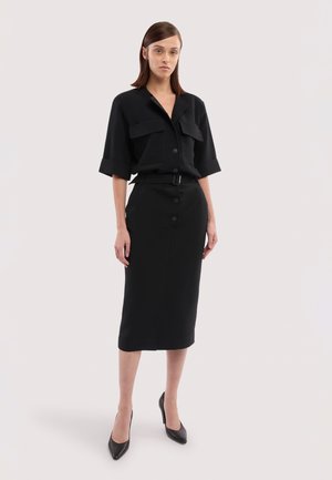 Femme debout portant une chemise noire à manches courtes avec boutons et poches, une jupe midi noire avec ceinture et des talons noirs.