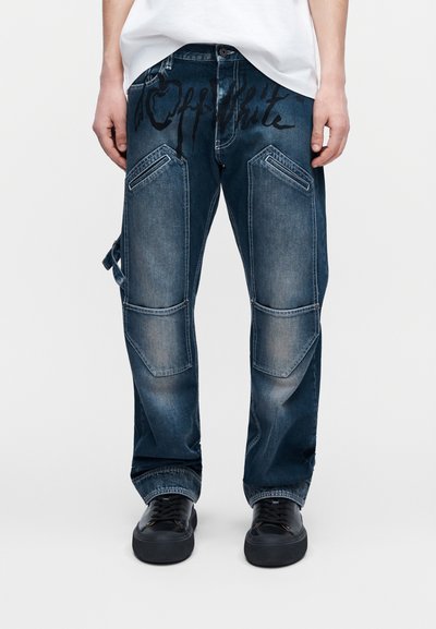 Jean en denim bleu foncé avec de grandes poches avant inclinées, renforts aux genoux décolorés, détail de sangle sur un côté, porté avec des baskets noires et une chemise blanche.
