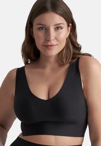 Ung kvinna med medellångt brunt hår iförd en svart V-ringad ärmlös crop top, som ser rakt fram med ett neutralt uttryck.