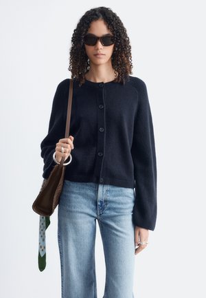 Femme aux cheveux bouclés portant des lunettes de soleil noires, un pull noir boutonné, un jean bleu clair, et portant un sac à main marron avec un foulard à motifs.