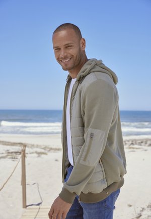 Hellgrüner Kapuzenpullover mit gepolsterter Vorderseite und langen Ärmeln, kombiniert mit einem weißen T-Shirt und blauen Jeans, vor einer Strandkulisse.