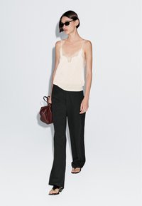 Lichte beige cami-top met kantafwerking, gecombineerd met hoge zwarte wijd uitlopende broeken. Houdt een kleine maroon tas vast. Zwarte sandalen.