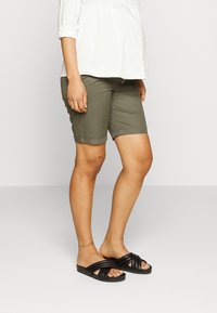Calções verde-azeitona com bainhas dobradas e detalhes de costura, combinados com sandálias pretas de escorregar com tiras cruzadas.