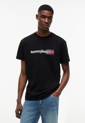 Άνδρας που φοράει μαύρο t-shirt Tommy Jeans και μπλε τζιν, στέκεται με το ένα χέρι στην τσέπη, μπροστά σε λευκό απλό φόντο.