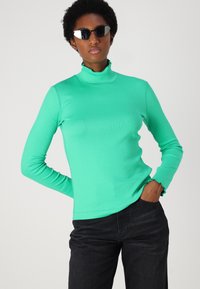 Mint green long-sleeve turtleneck top in ribbed fabric, featuring a ruffled collar and cuffs, paired with dark denim pants.