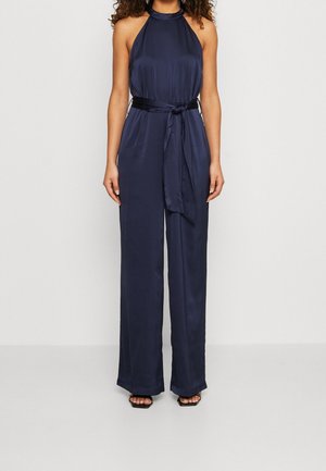 Donna che indossa una tuta smanicata blu navy con collo halter, pantaloni ampi e cintura annodata in vita, in piedi davanti a uno sfondo semplice.