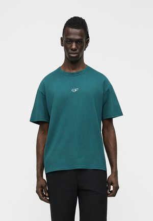 Άνδρας με σκουρόχρωμο δέρμα και κοντά πλεξούδες φοράει πετρόλ φαρδιά μπλούζα με μικρό λευκό λογότυπο και μαύρο παντελόνι, στέκεται μπροστά σε απλό φόντο.