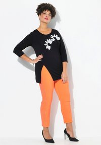 Tunic nera con applicazioni floreali bianche, abbinata a pantaloni stretti arancioni brillanti e scarpe con tacco nere. Design semplice e moderno.