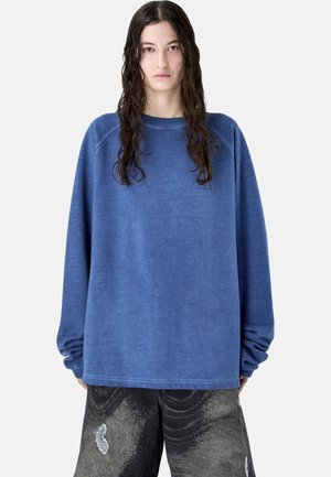 Młoda osoba z długimi mokrymi włosami, ubrana w za dużą, wyblakłą niebieską bluzę oraz luźne, czarne spodenki w wzory, stojąca na białym tle.