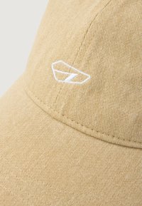 Gorra de pana beige con visera curva, que presenta un logotipo bordado en blanco en el frente y líneas texturizadas sutiles en toda su superficie.