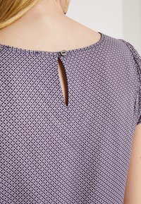 Lila mönstrad topp med rund halsringning, korta ärmar och en nyckelhålsdetalj bak, stängd med en knapp.