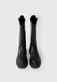 Une paire de bottes noires en cuir, hautes et avec des semelles épaisses, tenant debout sur un fond blanc uni.