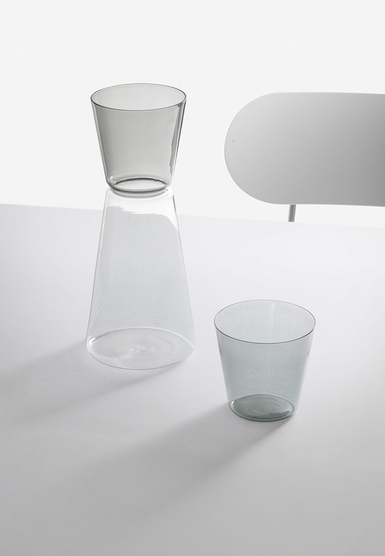 Przezroczysta szklana karafka z stożkową górą oraz pasującą szklanką. Oba elementy mają gładkie powierzchnie i subtelny szary odcień. Minimalistyczny design, bez wzorów.