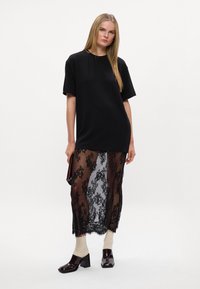 Czarny, oversize T-shirt nałożony na przezroczystą, czarną koronkową spódnicę z kwiatowym wzorem i falbaniastymi brzegami. Modelka ma na sobie ecru skarpetki i lakierowane szpilki.