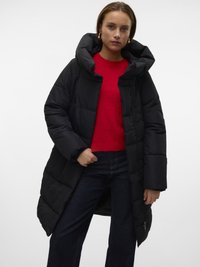 Nő fekete hosszú steppelt kabátban nagy kapucnival, piros pulóverben, sötét farmerben és arany karika fülbevalókkal, sima világos háttér előtt.