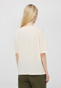 Beżowa bawełniana koszulka z luźnym krojem, krótkimi rękawami i okrągłym dekoltem, widoczna z tyłu. Prosty design, bez widocznych wzorów.