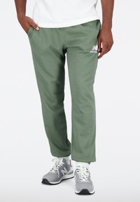 Zelené tepláky z měkké látky, s bočními kapsami a vyšitým bílým logem. Kombinovány se šedými sportovními botami.