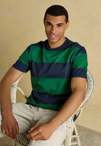Homme portant une chemise à manches courtes à rayures vertes et bleu marine et un pantalon beige, assis sur une chaise en osier blanche contre un mur beige.