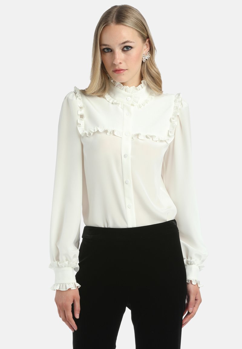 Bluză albă cu guler și manșete volănoi, având închidere cu nasturi pe față. Material neted, mâneci lungi și croială ajustată. Asortată cu pantaloni negri.