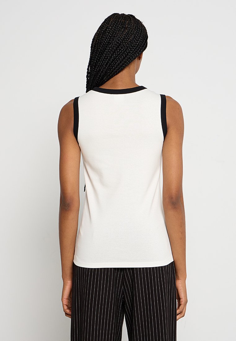 Biała bezrękawnik z czarnym wykończeniem przy dekolcie i otworach na ramiona, dopasowany krój, w zestawieniu z pasiastymi spodniami w czarno-białym kolorze.