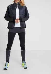 Personne portant une veste matelassée noire, un pantalon de sport noir et gris, un haut blanc, et des chaussures de course bleu clair et jaune néon, debout les mains jointes.