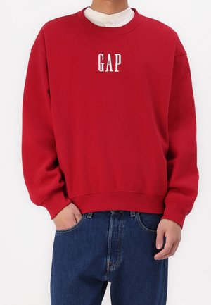 Person iført en rød sweatshirt med hvidt GAP-logo, lagt over en hvid skjorte og blå jeans med den ene hånd i lommen.