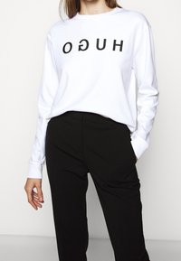 Witte sweatshirt met lange mouwen en zwarte "HUGO" tekst die ondersteboven is gedrukt, gecombineerd met zwarte broek. Eenvoudig, ontspannen ontwerp en pasvorm.
