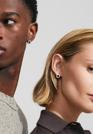 Studs met een zwarte steen in het midden, omringd door heldere kristallen; weergegeven aan het oor van een model. Matte en gepolijste texturen zijn duidelijk zichtbaar.