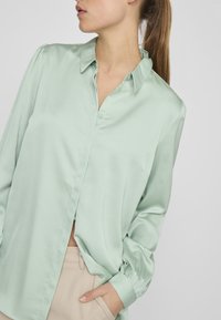 Light green satin long-sleeve shirt with a collar, button front, and gathered detailing at shoulders. Paired with beige pants.