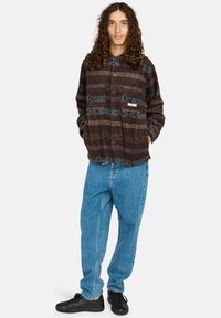 Giovane con capelli lunghi e ricci, che indossa una giacca marrone a fantasia, jeans blu larghi e sneakers nere, in piedi con le mani in tasca.