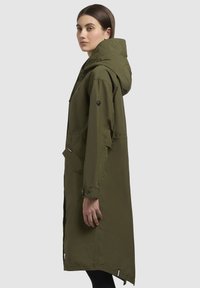 Manteau de pluie long vert olive avec capuche, poches latérales et poignets boutonnés. Texture lisse et design droit.