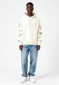 Sudadera crema con un bolsillo tipo canguro, que presenta gráficos multicolores en las mangas. Combinada con jeans azul claro y zapatillas blancas.