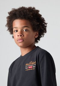 Juodas medvilnės marškinėlis su apvalia iškirpte, puošnus daugiarūšiu siuvinėtu logotipu ant krūtinės. Modelis turi trumpus, garbanotus plaukus.