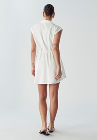 Vestido blanco sin mangas con cuello tipo polo y cintura con cordón, que presenta una falda acampanada y una textura suave. La vista trasera muestra un diseño simple.