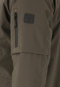 Olivgrön jacka med en slät textur, med en ficka med dragkedja på vänster ärm och en svart logotypapplikation.