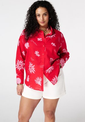 Femme aux cheveux bouclés portant une chemise rouge à boutons avec des imprimés de créatures marines roses et un short blanc, debout avec une main dans la poche.