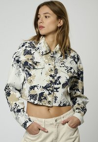 Chaqueta de mezclilla recortada con un patrón de camuflaje en beige, marino y blanco. Tiene dos bolsillos en el pecho y un cierre de botones.