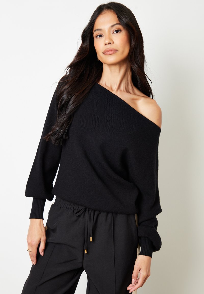 Zwarte off-the-shoulder trui met lange mouwen, gemaakt van zachte stof. Gecombineerd met zwarte joggingbroek met een trekkoord en gouden accenten.