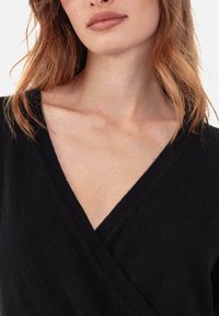 Pull noir à col en V en tissu doux, présentant un design enveloppant et un tricot texturé. Gros plan sur l'encolure et le haut du torse avec des détails minimaux.