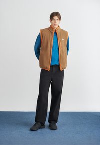 Hnědá fleece vesta s oranžovými detaily, zapínáním na zip, nošena přes modrou košili s dlouhým rukávem. Černé kalhoty širokého střihu a černé boty doplňují outfit.