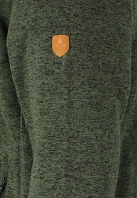 Tessuto di un verde scuro e textured con una piccola etichetta in pelle marrone, etichettata "Whistler", cucita nella zona del petto.