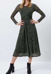 Langärmliges, figurbetontes grünes Kleid mit glitzernder Textur, geraffter Taille und schwingendem Rock. Getragen mit schwarzen Ankle Boots.