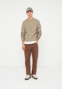 Maglione di lana marrone, sottoshirt chiaro, pantaloni marroni e sneakers beige. Accessoriato con un berretto chiaro. Outfit semplice e casual.