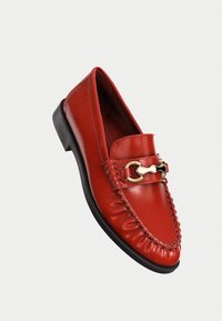 Red leather loafers with a rounded toe, stitched detail along the front, and a gold-tone hardware accent on the strap. Flat black sole.