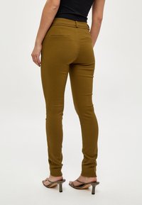 Olivgröna figurnära byxor med en slät textur och två bakfickor, tillsammans med ljusfärgade högklackade sandaler.