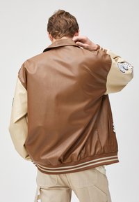 Bruine faux leren jas met beige mouwen, ribbelmanchetten en onderkant. Heeft een geTEXTUREERD afwerking en geborduurde details op de mouw.