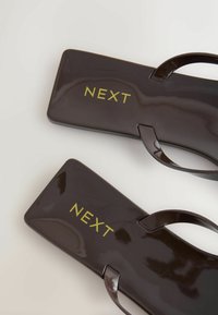 Sötétbarna, téglalap alakú papucsok négyzet alakú orral és vékony pántokkal, amelyeken a "NEXT" szó sárga színnel van nyomtatva mindkét talpon.