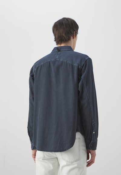 Marineblå denimskjorte med lange ermer, krage og knapper ved mansjettene. Har en bakyoke og en opphenget for nakken.