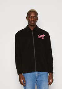 Giacca in pile nera con chiusura a zip, colletto e logo ricamato rosso sul lato sinistro del petto. Indossata con jeans azzurri chiari.